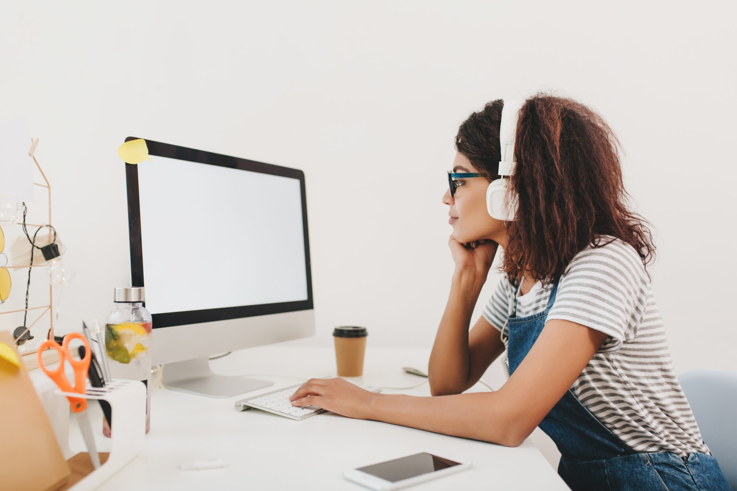 Slim black girl in headphones looking at computer's screen with attention. Indoor portrait of young curly woman doing her work in office and drink coffee.